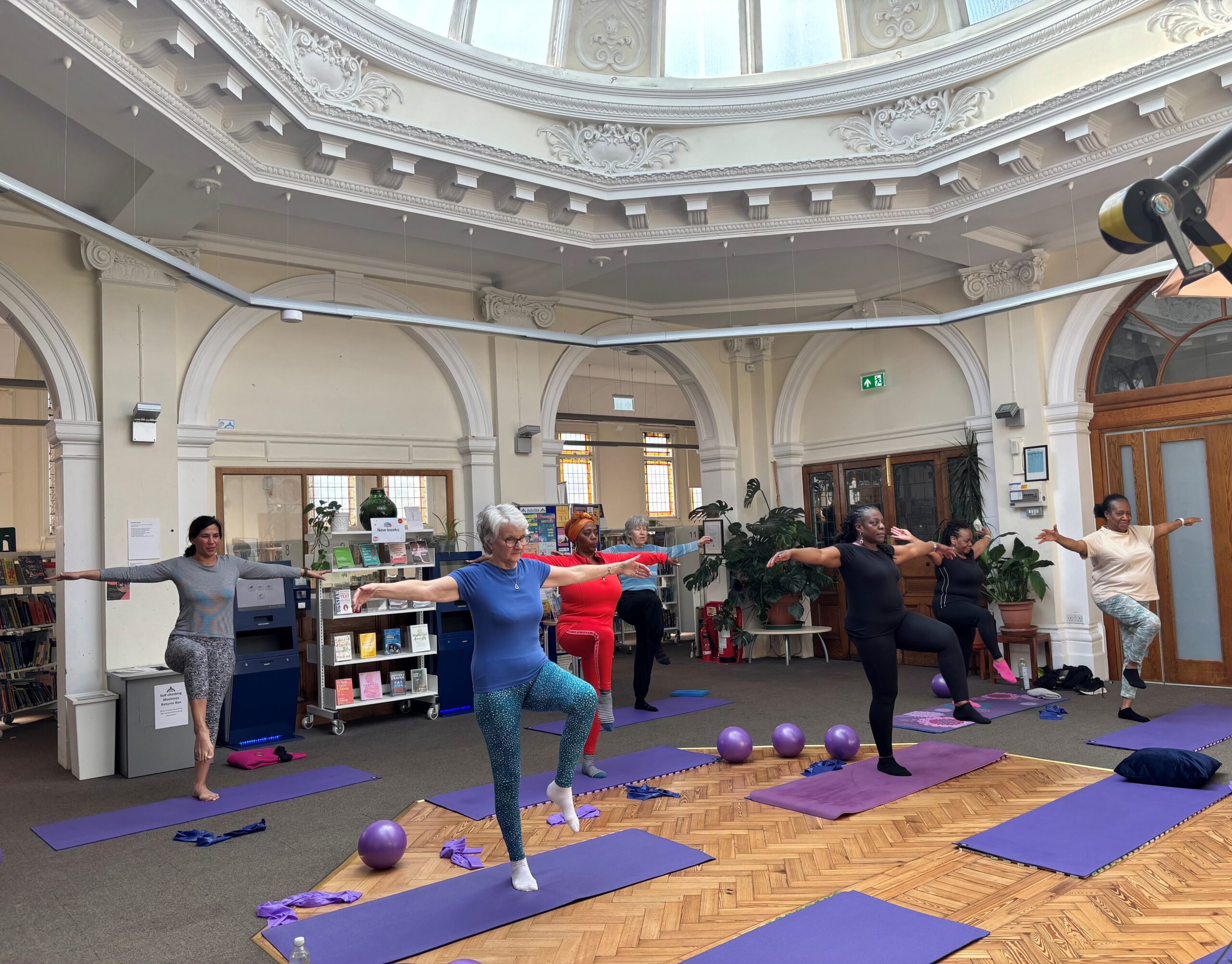 Group fitness class doing yoga poses on purple mats in ornate library with domed ceiling and classical architecture