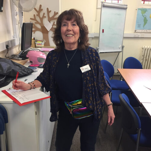 Teacher with brown hair smiling in classroom, wearing dark cardigan and holding colorful supplies at desk