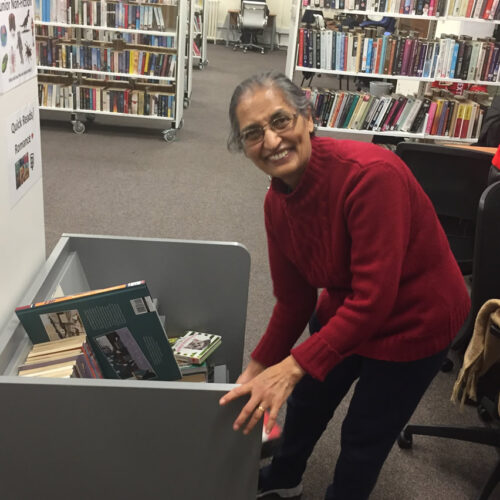 Smiling woman in red sweater organizing books in library cart surrounded by bookshelves and reading area