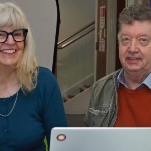 Two professionals collaborating at a white desk with laptop, documents, and office supplies in modern workplace setting.