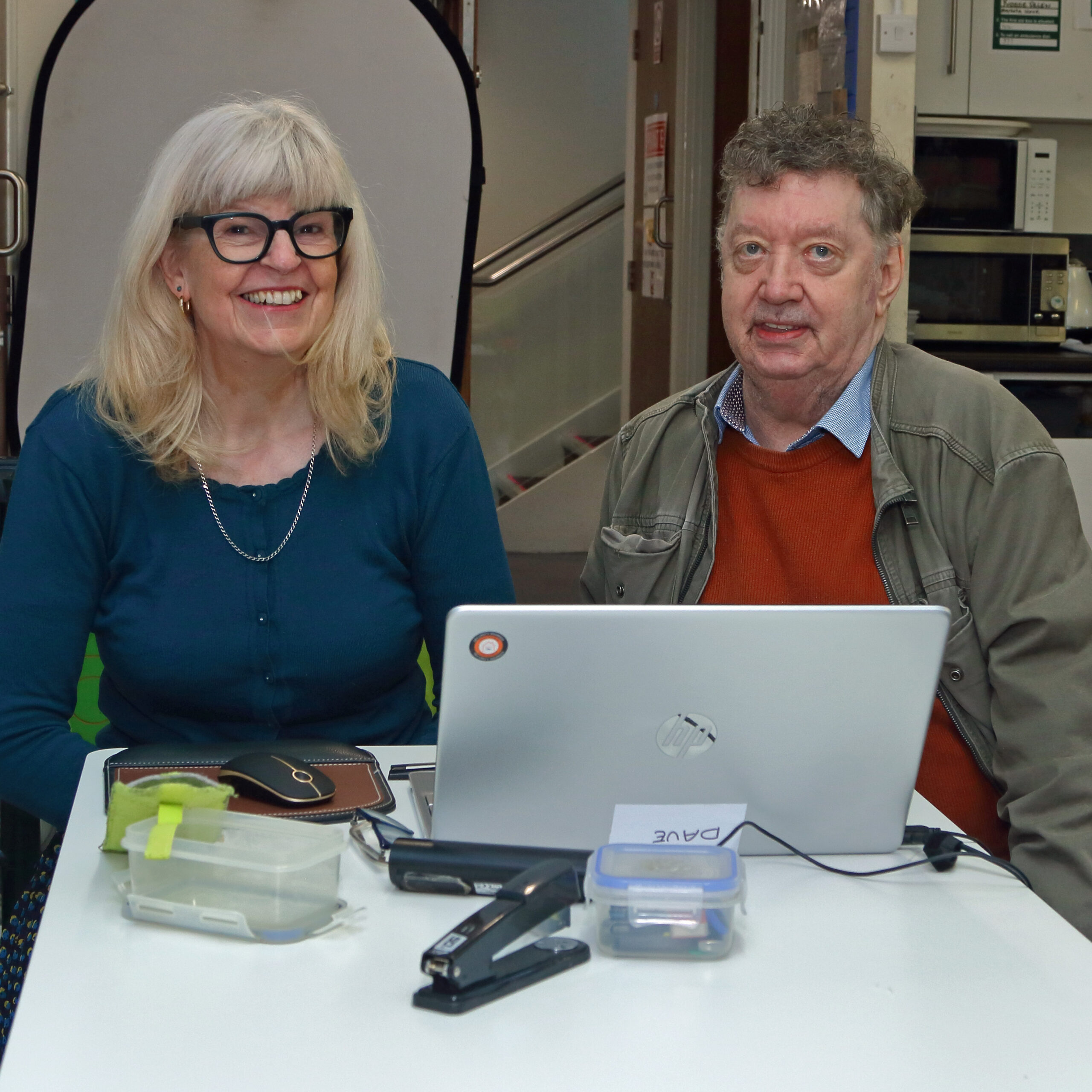 Two professionals collaborating at a white desk with laptop, documents, and office supplies in modern workplace setting.