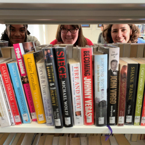 Three smiling women browse books on library shelf, with various book titles visible including biographies and fiction