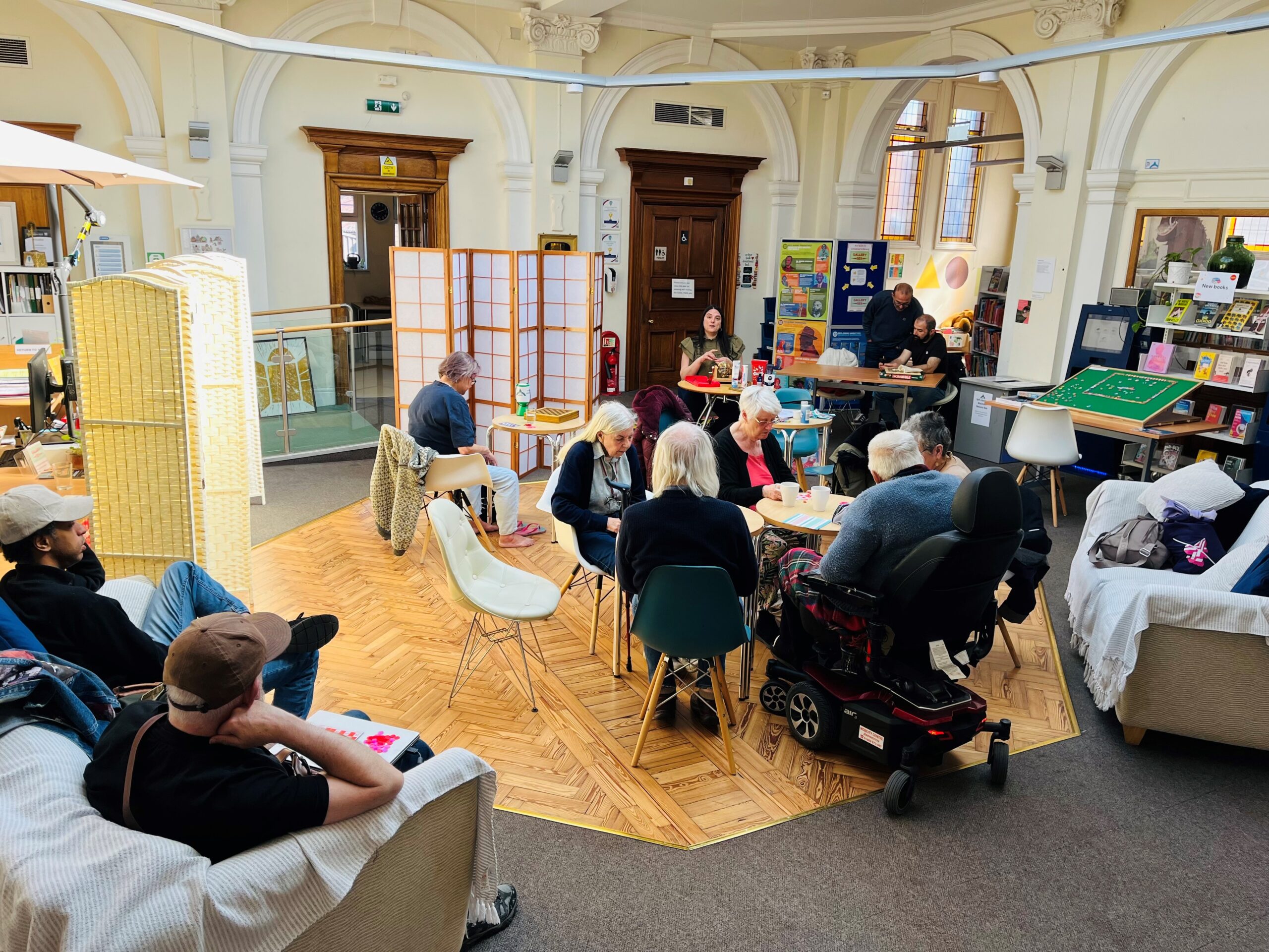 People of various ages gather in a historic library with arched architecture, some seated at tables, others on sofas reading.