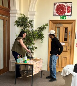 Woman in olive vest serving hot beverage from table to man in white cap and black sweater in elegant interior room