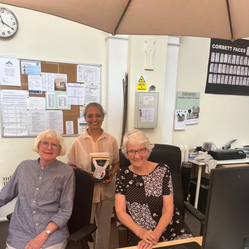 Three smiling women in office setting, one holding award plaque, with bulletin boards and desk visible in background.