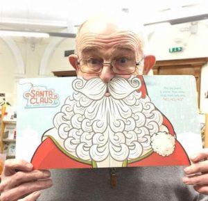 Elderly man holding Santa Claus face cutout with white beard and red hat in indoor setting