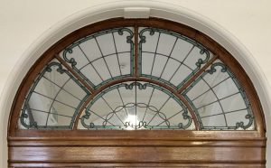 Arched stained glass window with ornate green scrollwork design and wooden frame in white archway
