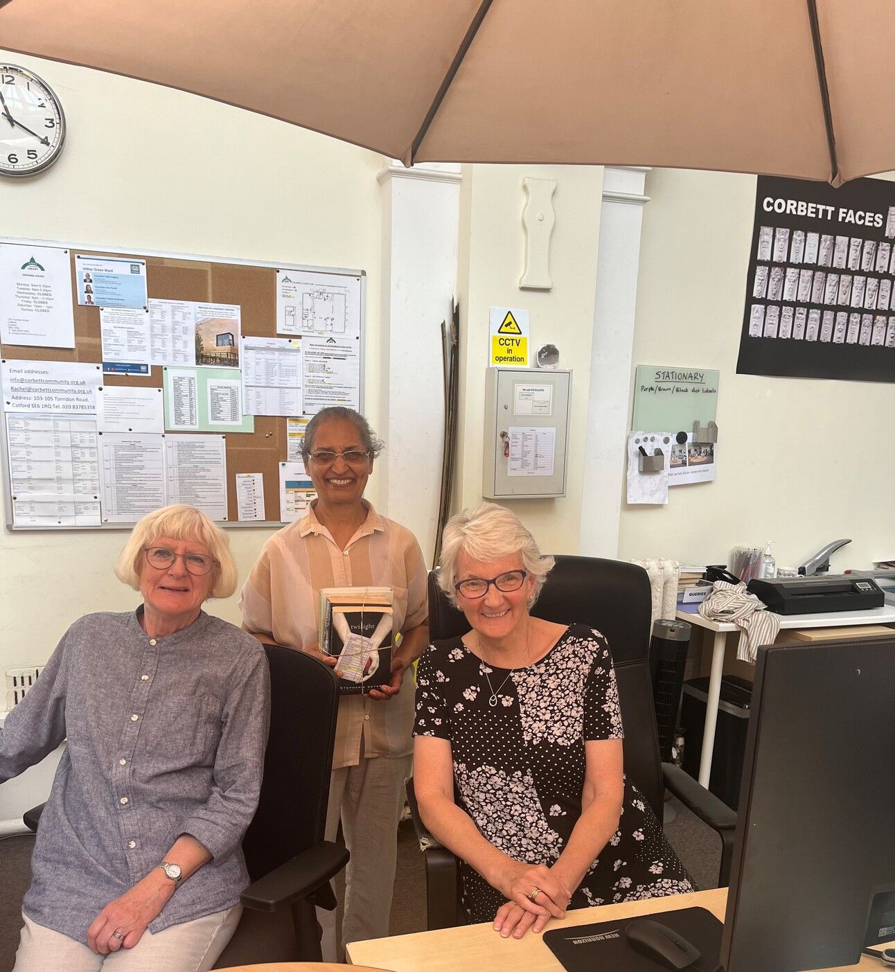 Three women in office setting, one holding award or plaque, with bulletin boards and desk visible in background.