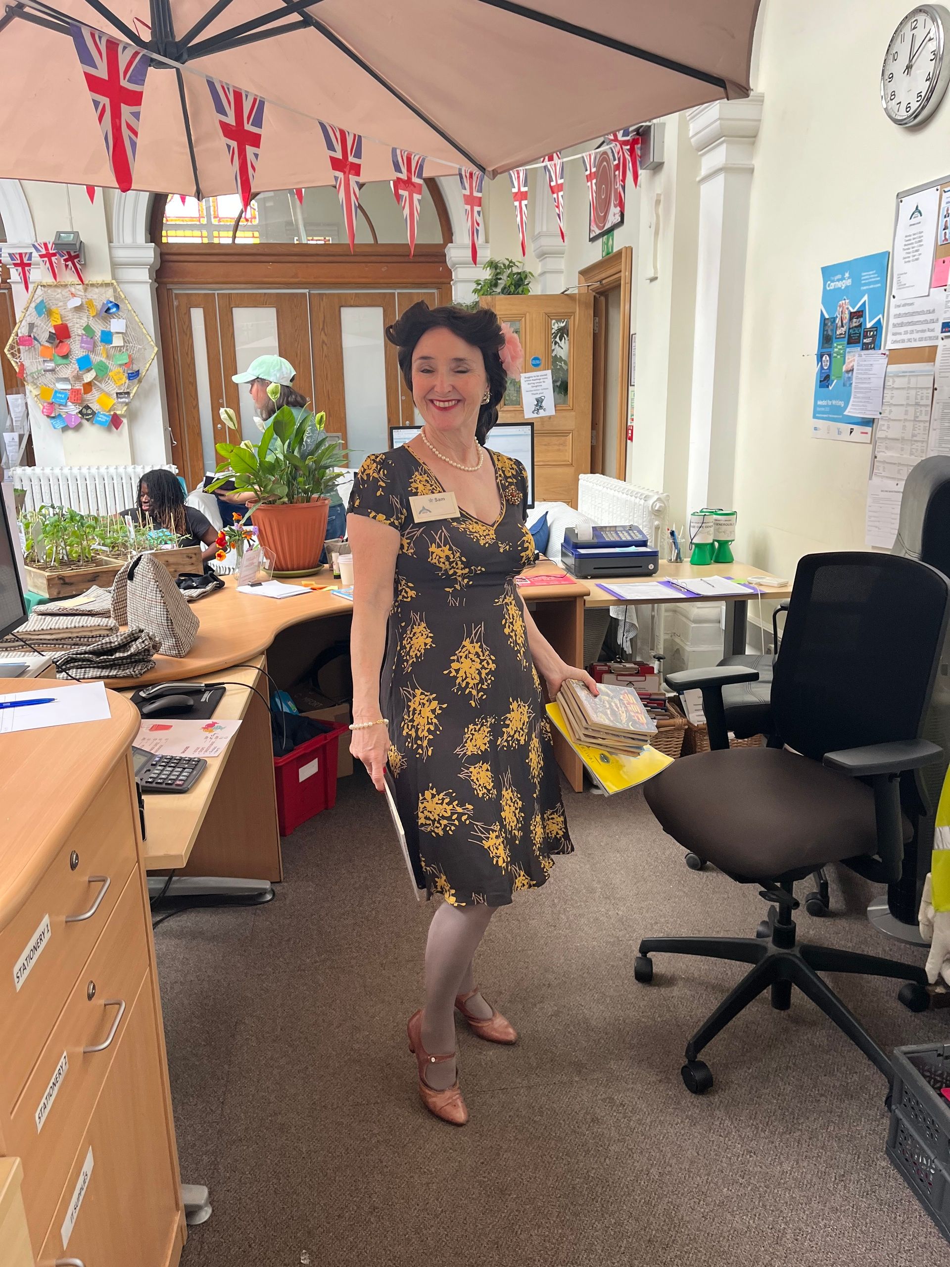 Woman in floral dress smiling in office decorated with British Union Jack bunting and plants on desks