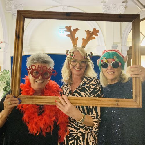 Three women in festive Christmas accessories posing with wooden picture frame in decorated library or office space