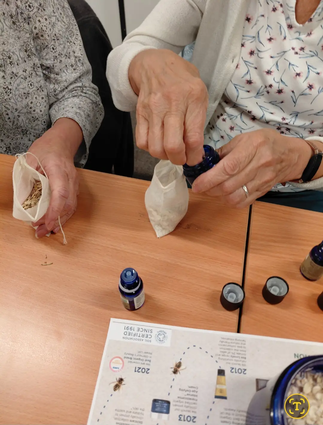 Elderly hands crafting with small bottles and white fabric pouches on wooden table during workshop activity