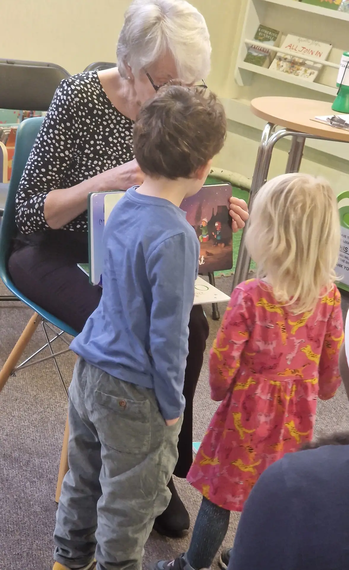 Grandmother reading picture book to two young children in library or classroom setting with bookshelves in background