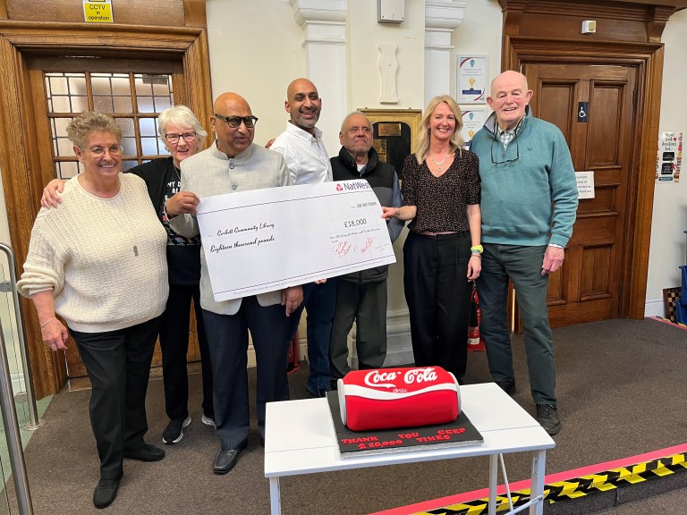 Group of people holding large £18,000 donation check for Cockfield Community Library with Coca-Cola cake on table