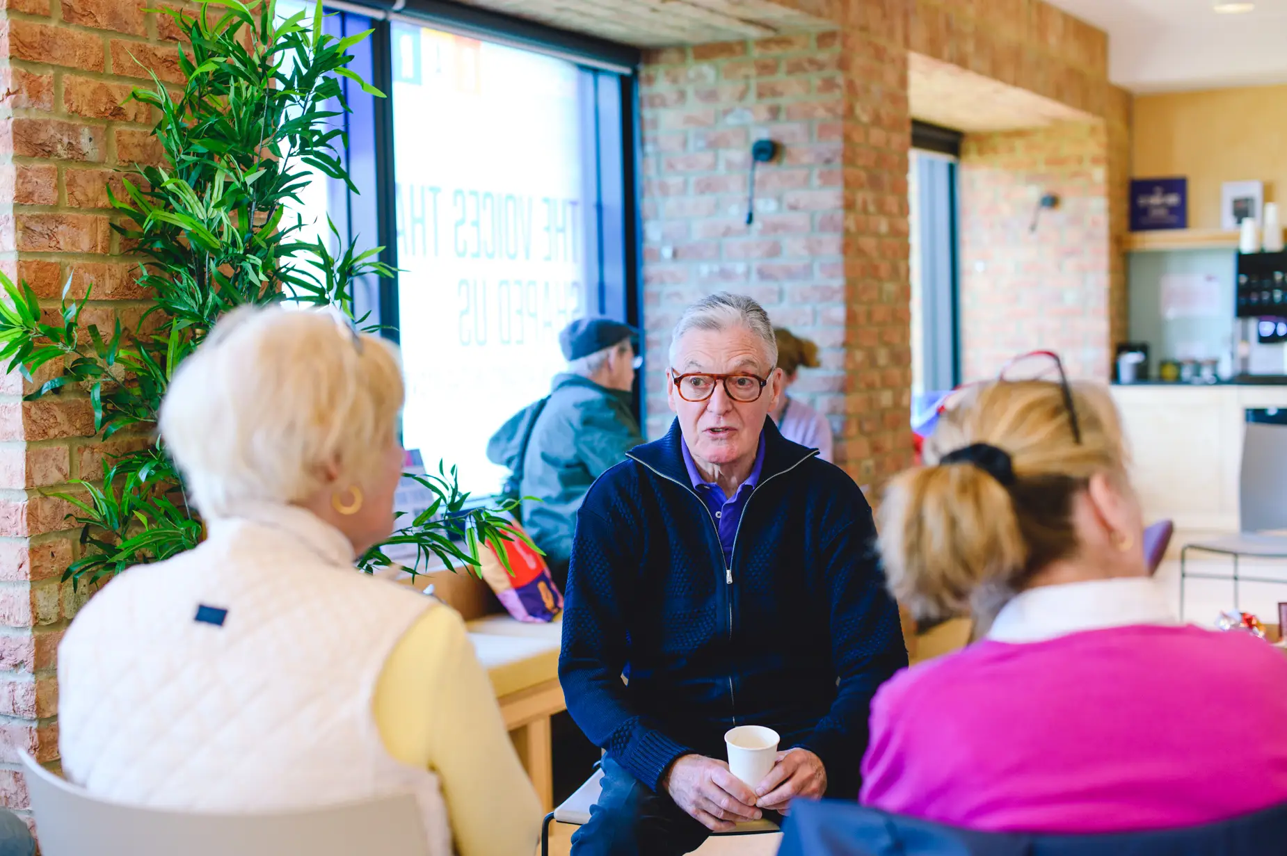 Elderly man with glasses talking to group of seniors in bright community center with brick walls and plants