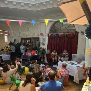 Musicians performing acoustic guitars for seated audience at indoor community event with colorful bunting decorations