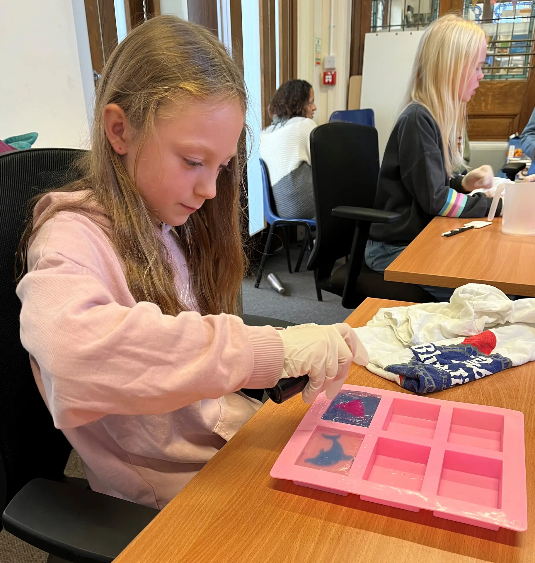 Young girl in pink sweatshirt doing printmaking art activity at wooden table in classroom setting