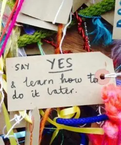 Handwritten tag reading Say YES & learn how to do it later surrounded by colorful yarn and ribbons.