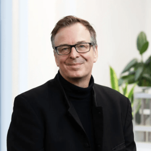 Smiling man with glasses wearing black turtleneck and blazer, professional headshot with plant in background.