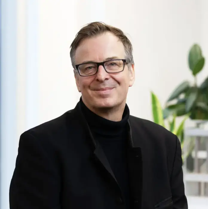 Smiling man with glasses wearing black turtleneck and blazer, professional headshot with plant in background.
