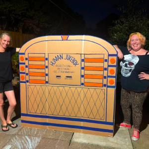 Two smiling women standing beside a large handmade cardboard Human Jukebox prop painted orange and blue in a garden
