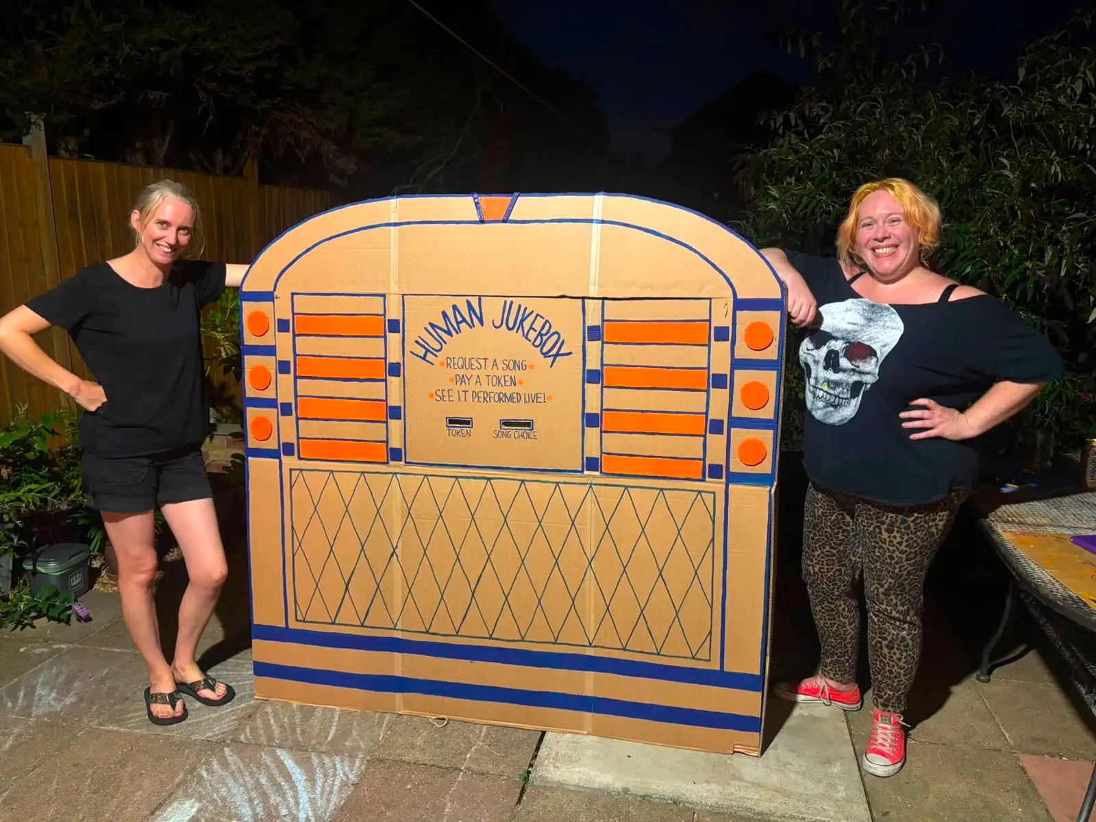 Two smiling women standing beside a large handmade cardboard Human Jukebox prop painted orange and blue in a garden