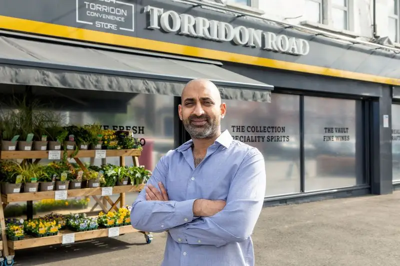 Smiling businessman with crossed arms standing outside Torridon Road convenience store with plant display