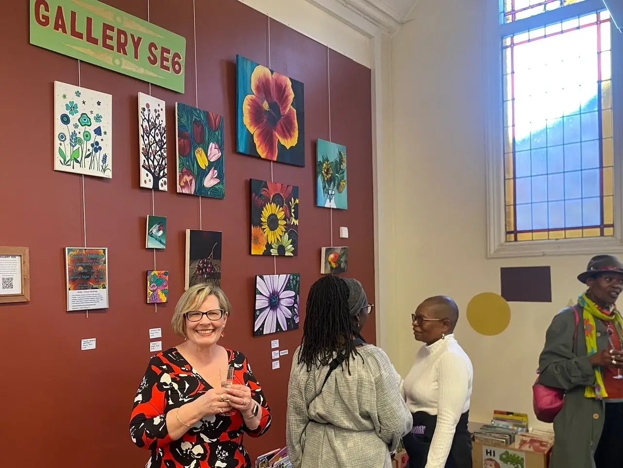 Visitors socializing at Gallery SE6 art exhibition, with colorful floral paintings displayed on a red gallery wall.