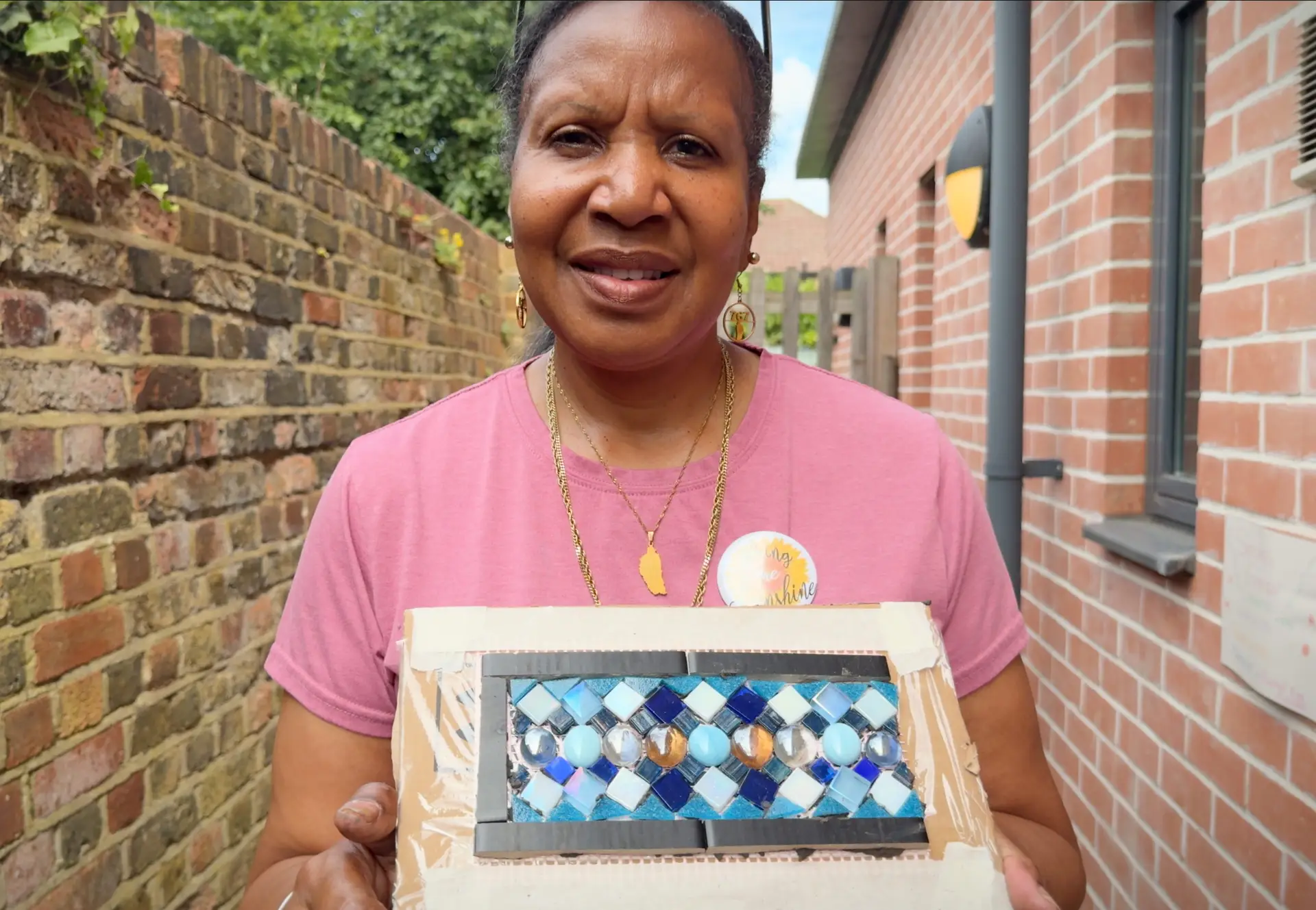 Woman in pink T-shirt with sunshine badge holding a handmade blue and white glass tile mosaic panel outdoors.