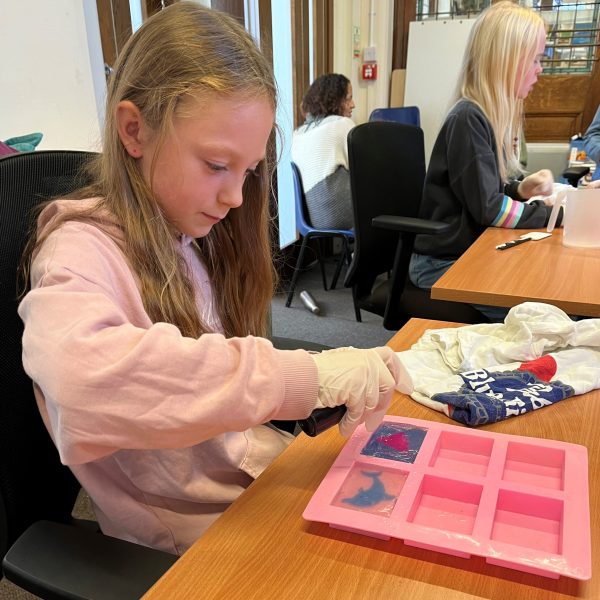 Girl in pink sweatshirt doing printmaking art project at wooden table in classroom setting