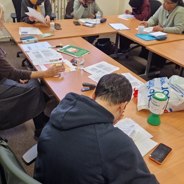 Group of adults studying and working together at tables with papers, books, and materials in classroom setting.