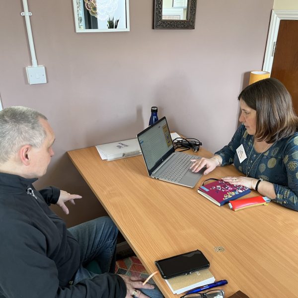 Woman in blue patterned shirt meeting with man at wooden desk with laptop and books in office setting