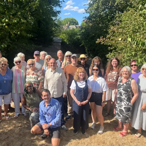Group of about twenty smiling volunteers posing together outdoors on a sunny day in a leafy garden.