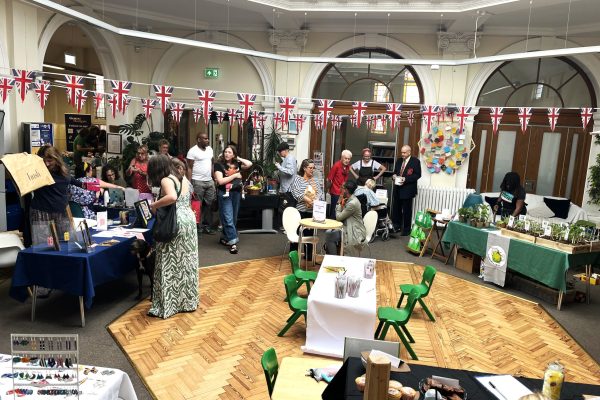 Community fair in historic building with British bunting, vendor stalls, families browsing crafts and local products