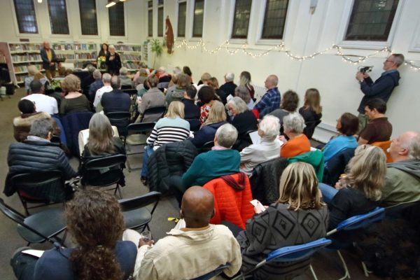 Audience seated in chairs listening to speakers at library event with string lights and bookshelves visible