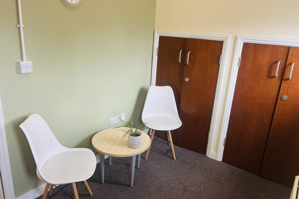 Modern office waiting area with white chairs, round table, plant, and wooden storage cabinets against green wall