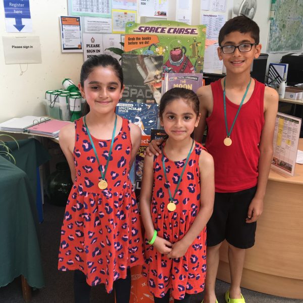Three children wearing medals pose in library with space-themed books and displays visible in background
