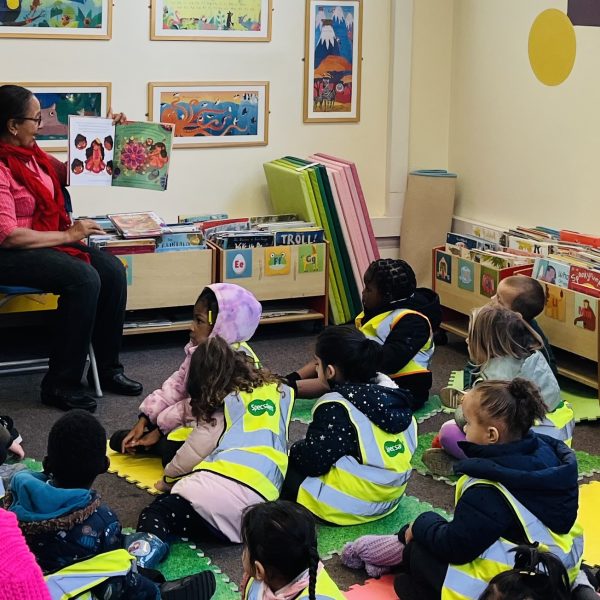 Teacher reading to diverse group of children in high-vis vests sitting on colorful mats in classroom library corner