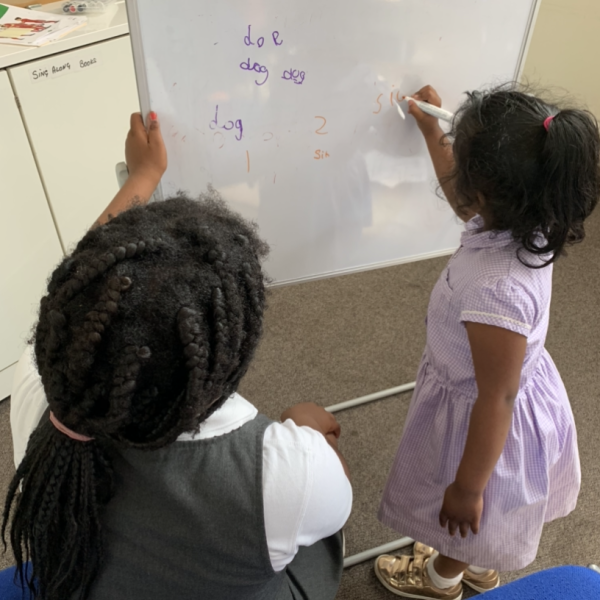 Two young students writing on whiteboard during classroom lesson, one in white shirt, one in purple striped dress